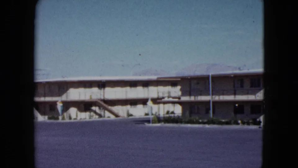 1959 Nevada 8mm frame of a low motel building, exterior corridor, stairs, lamp post, and empty foreground pavement.
