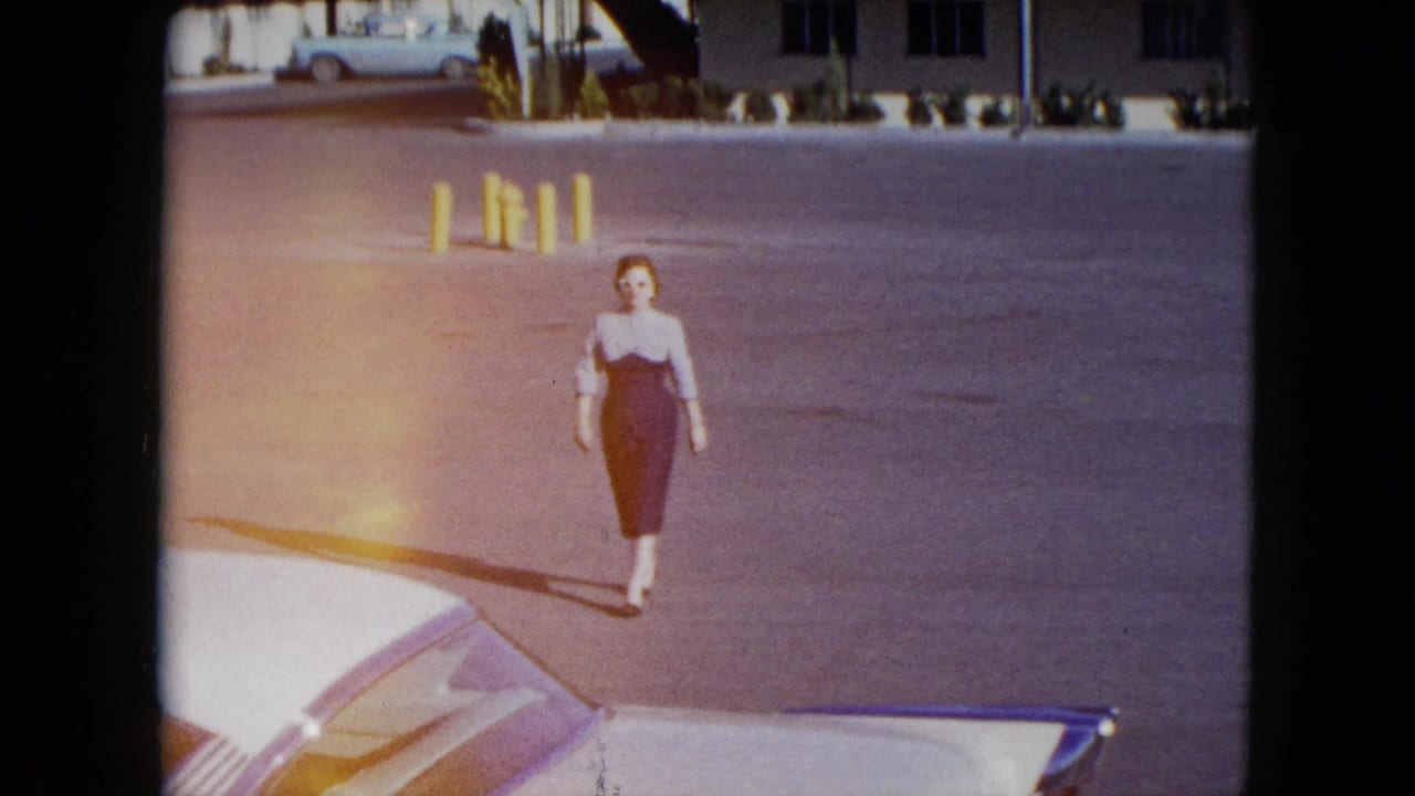 In 1959 Nevada home-movie footage, a woman in a dark and pale fitted dress walks across a parking lot toward a white car.