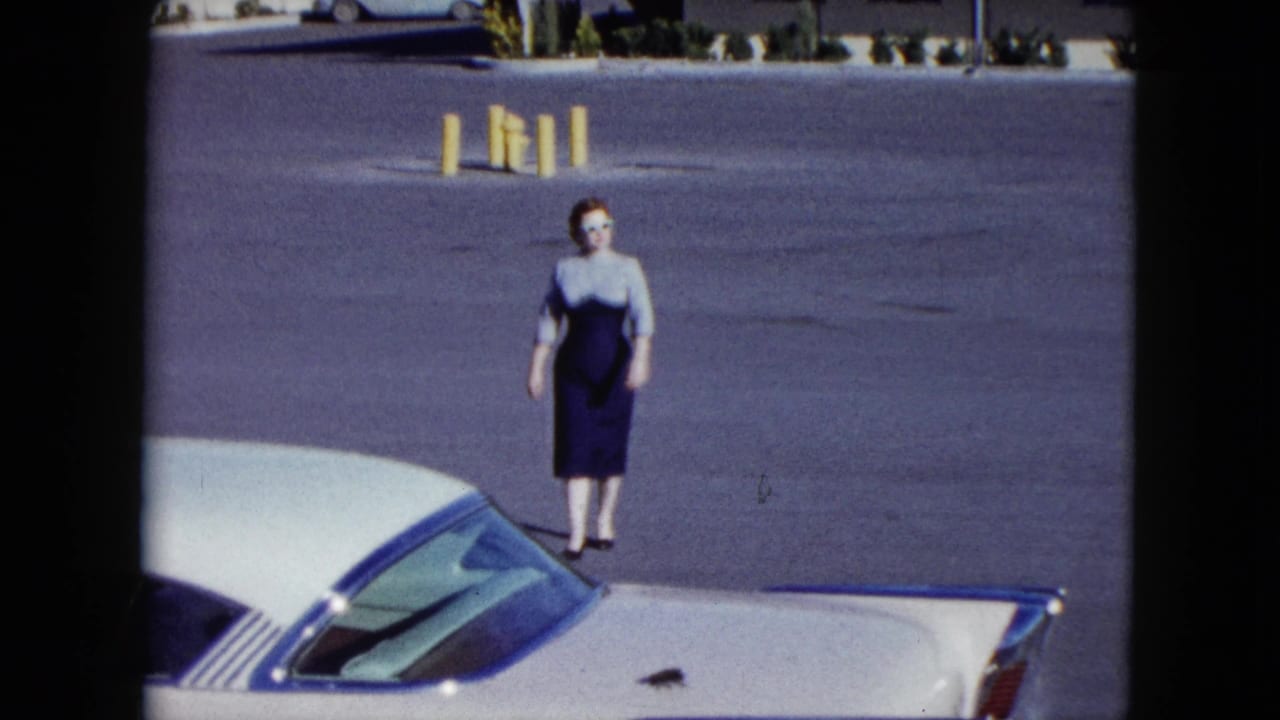 Woman stands or walks near the front of a white car in a broad asphalt lot.