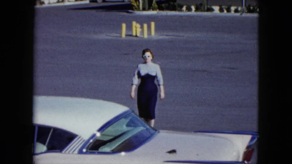 The woman in sunglasses and a fitted dress passes beside the white car, framed by asphalt and yellow parking bollards.