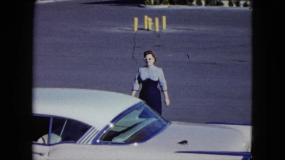 At the end of the clip, the woman stands near the side of the white car, with the sunlit parking lot and bollards behind her.