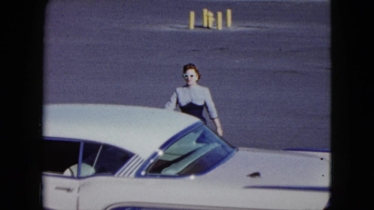 Woman in a 1950s-style dress standing beside a white car near a wall.