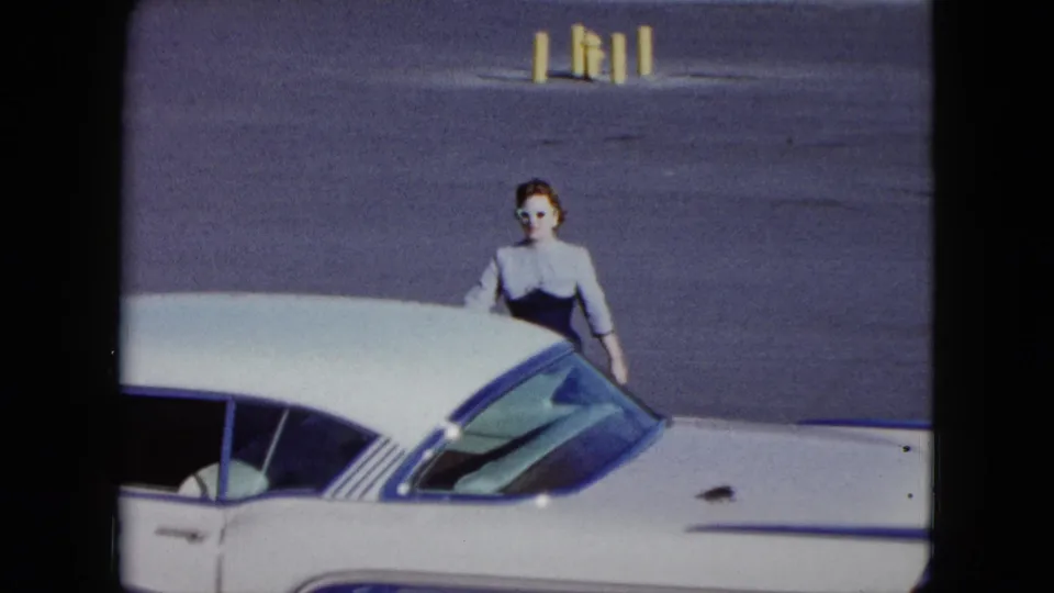 In metadata-identified 1959 Nevada footage, a woman stands beside a white classic car with blue trim in a posed travel shot.