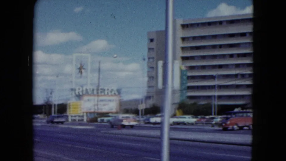 The handheld 8mm view frames the Riviera sign, multi-story hotel building, street lanes, and period automobiles in Nevada.