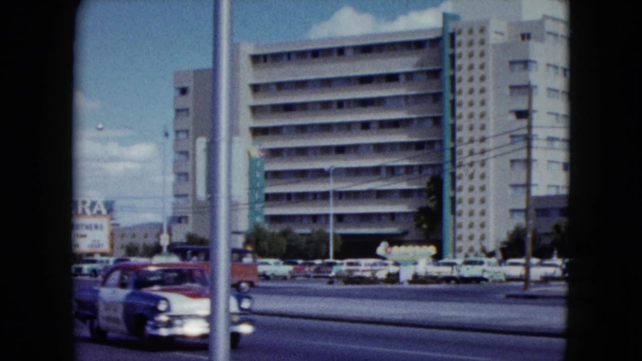 Classic cars pass a large hotel building with street poles in the foreground.