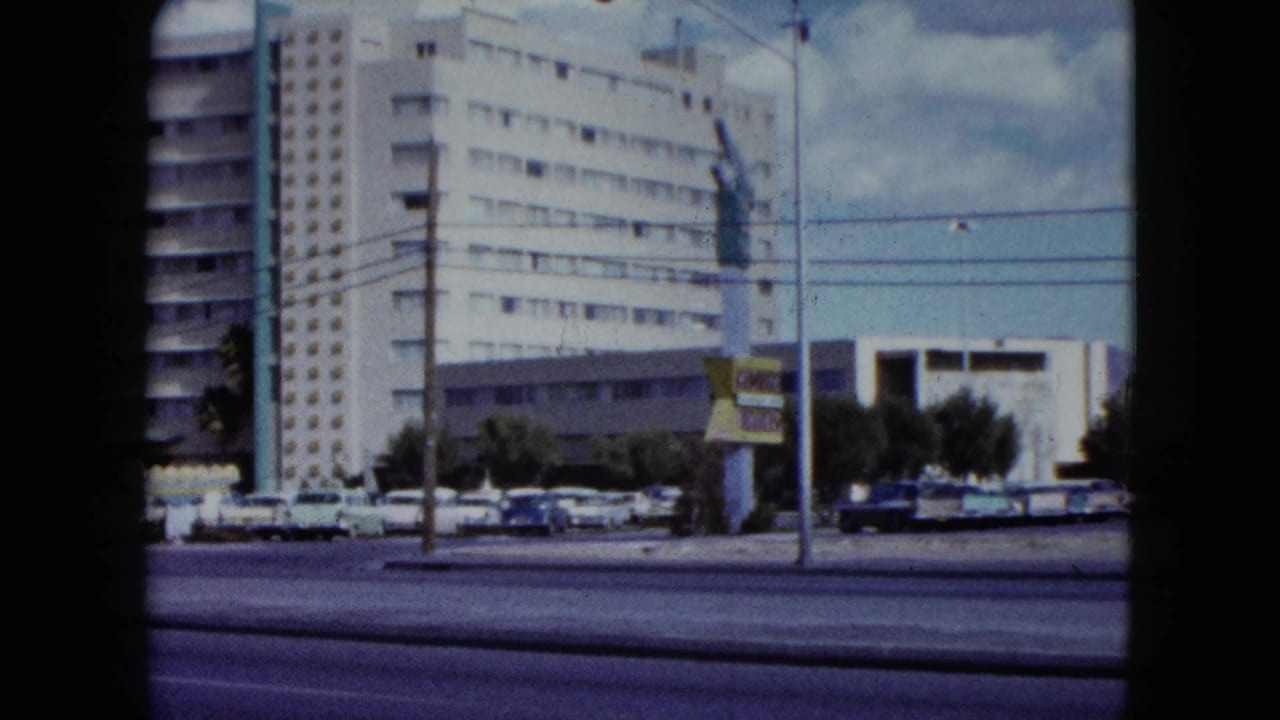 Street with moving cars, parked cars, utility lines, and a tall modern building.