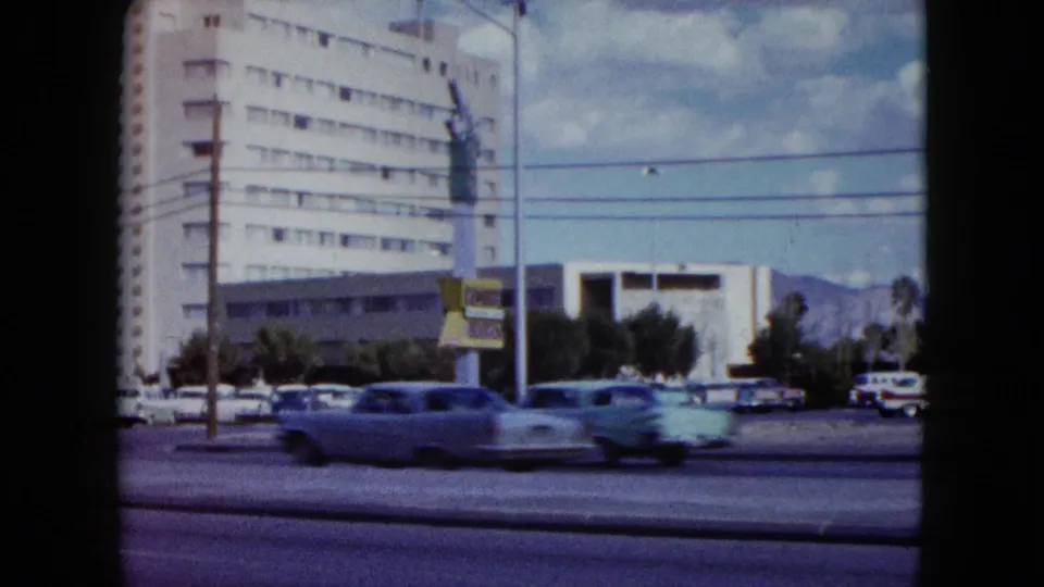 Mid-century cars move along a Nevada street as the camera pans past a modern building, parking area, wires, and distant mountains.
