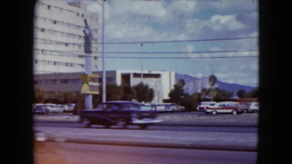 A blurred car crosses the foreground during the 1959 Nevada pan, with parked vehicles, low buildings, utility lines, and mountains behind.