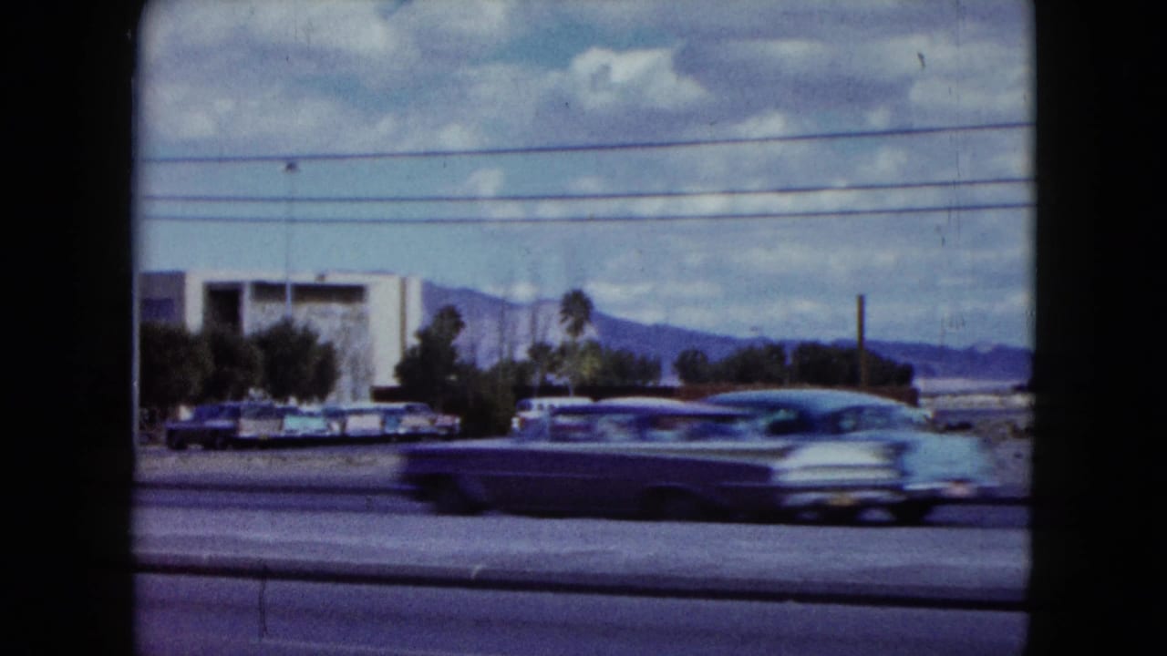 1959 Nevada road-view frame with a classic car passing low roadside buildings, utility wires, cloudy sky, and distant mountains.