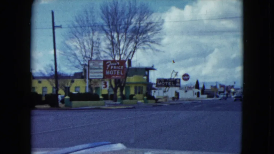 1959 Nevada roadside view showing a motel sign, broad street, utility pole, low buildings, leafless tree, cloudy sky, and mountains.