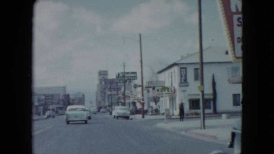 1959 Nevada passing street view with roadside motel signs, moving cars, sidewalks, and small pedestrian figures.