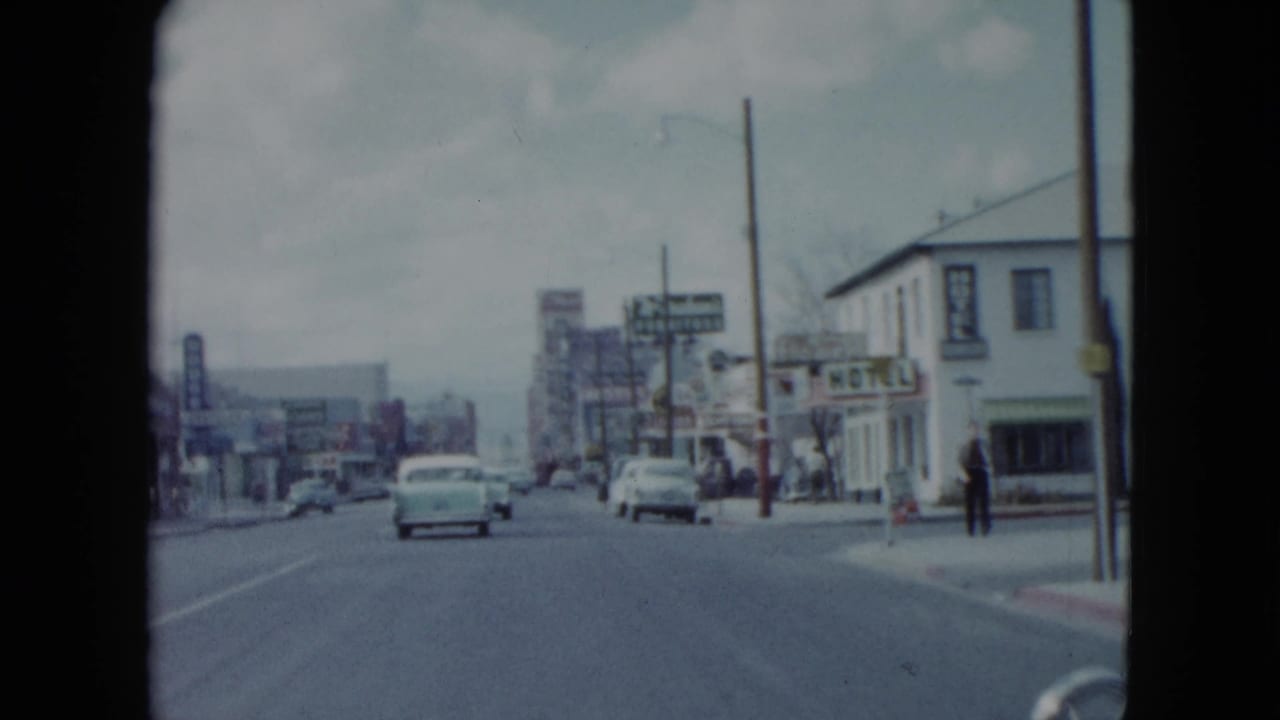 1959 Nevada street scene from a moving car, showing mid-century automobiles, storefront signs, utility poles, and pedestrians on the sidewalk.