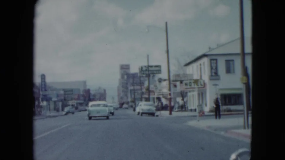 1959 Nevada street scene from a moving car, showing mid-century automobiles, storefront signs, utility poles, and pedestrians on the sidewalk.