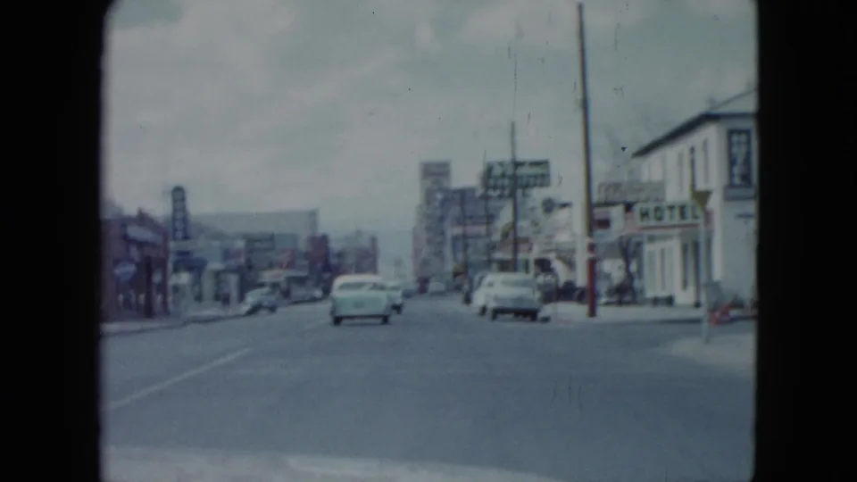 A moving-vehicle view of a 1959 Nevada commercial street with classic cars, shopfronts, signs, and a hazy overcast background.