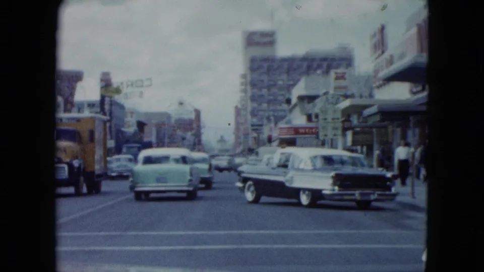 1959 Nevada downtown traffic with a pale car ahead, a yellow delivery truck, a two-tone car turning, pedestrians, storefront awnings, and a hotel sign.