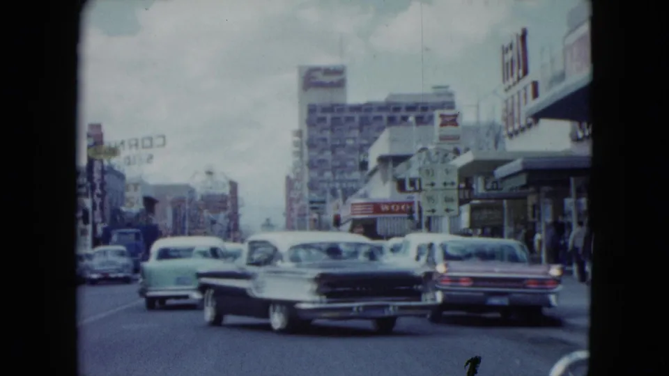 1959 Nevada street traffic seen from a moving car, with multiple mid-century automobiles, storefronts, pedestrians, and prominent commercial signage.