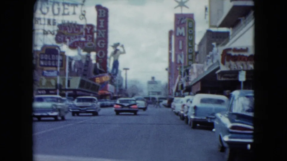 1959 Nevada traffic scene with classic cars, vertical neon signs, casino frontage, and sidewalk pedestrians in the distance.