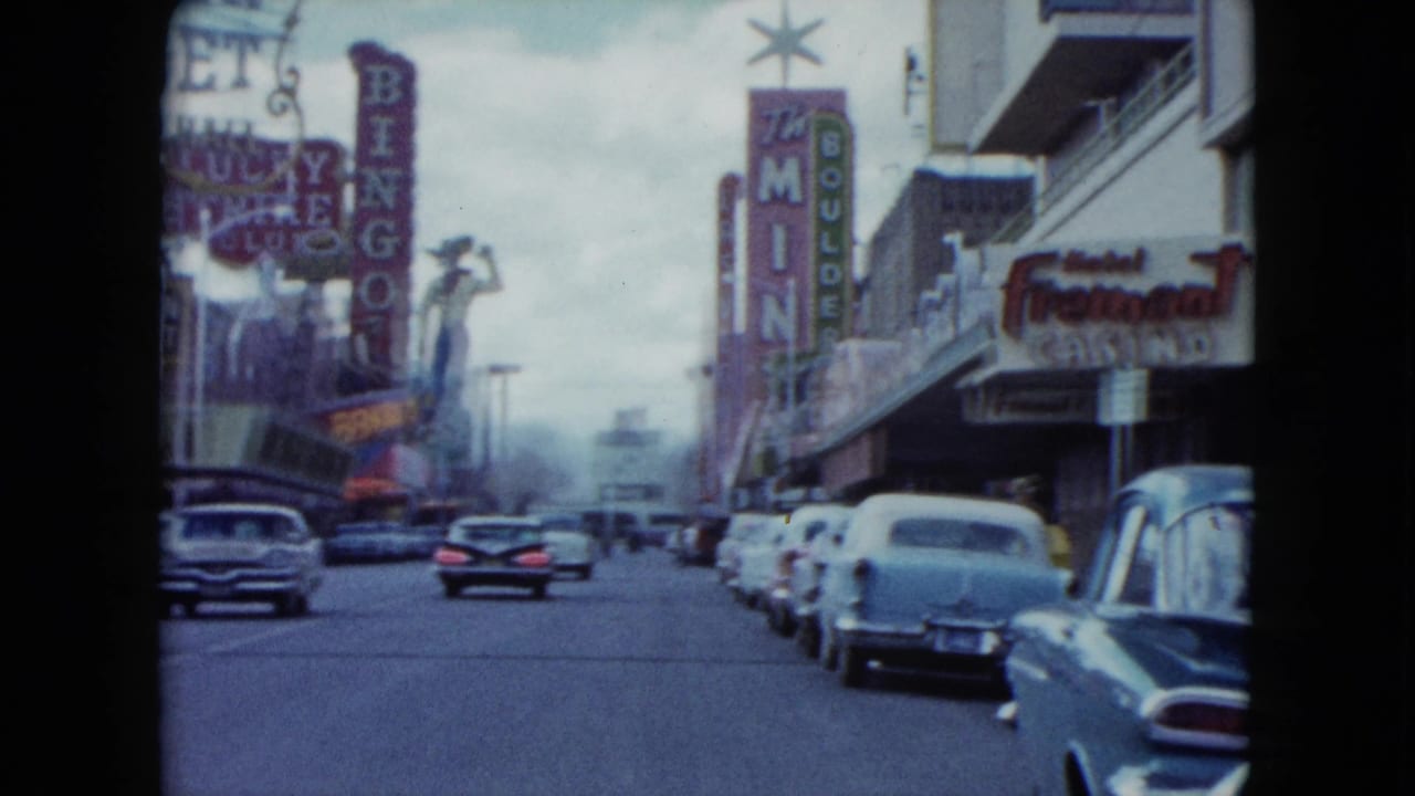 1959 Nevada home-movie view down a casino street with classic cars, vertical neon signs, and storefront marquees.