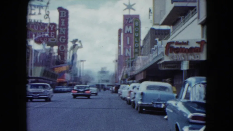 1959 Nevada home-movie view down a casino street with classic cars, vertical neon signs, and storefront marquees.