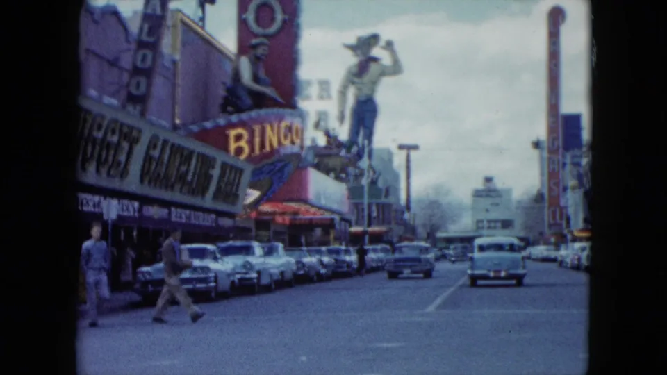 1959 Nevada street scene with parked classic cars, pedestrians, casino and bingo signage, and a large cowboy figure above the roadway.