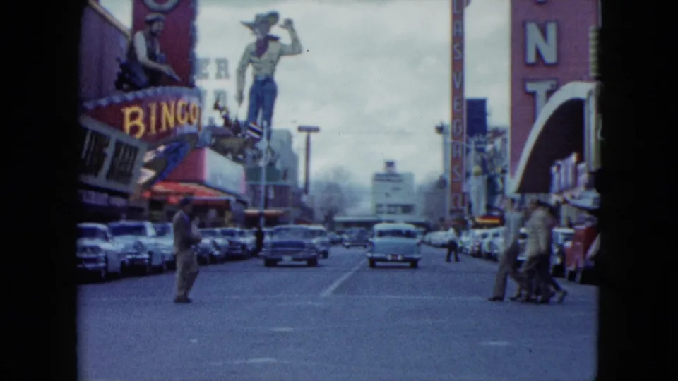 1959 Nevada downtown view with cars parked along both sides, people crossing the road, casino signage, and a cowboy statue near a bingo hall.