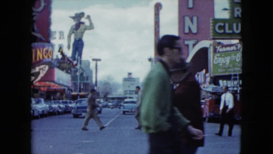 1959 Nevada street footage with two pedestrians close to camera, parked cars, a bingo sign, casino signage, and a cowboy statue beyond them.