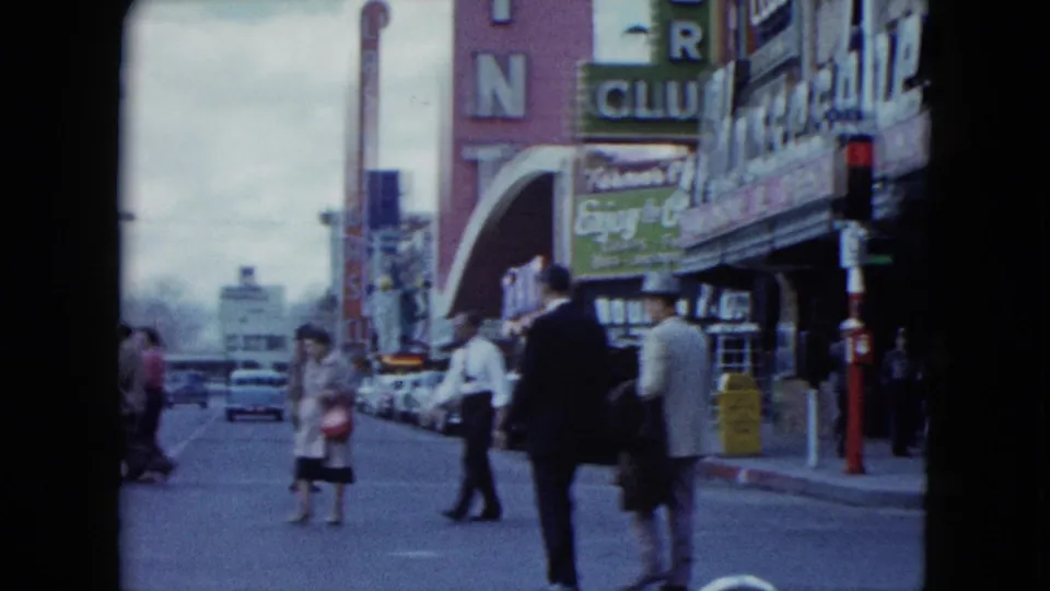 1959 Nevada commercial street with pedestrians near a casino frontage, a green entertainment sign, parked cars, and distant traffic.
