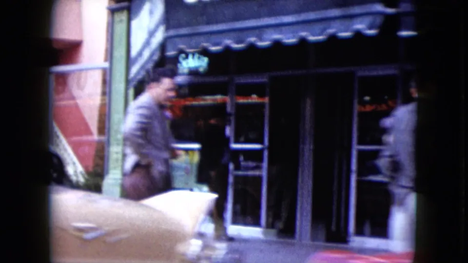 1959 Nevada street view with a suited pedestrian, glass storefronts, awning, and a classic car moving through the foreground.