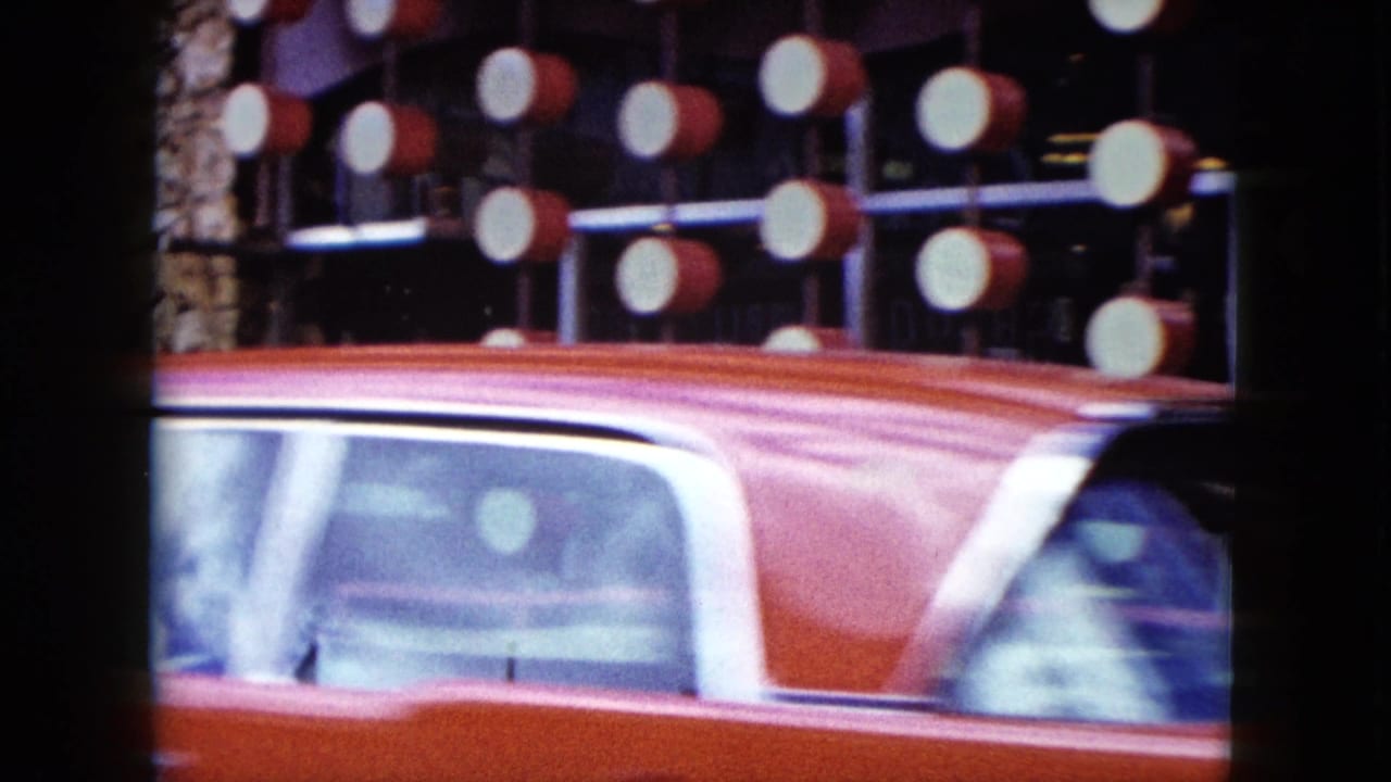 Pink 1950s car passing illuminated storefront lights on a city street.