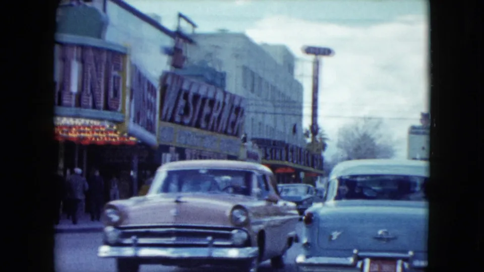 Two mid-century cars pass a Nevada commercial block with gambling hall lettering and Western-themed storefront signage.
