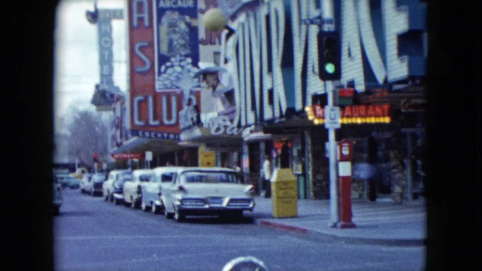 1959 Nevada downtown corner showing parked classic cars, neon casino signage, a traffic signal, and pedestrians near storefronts.