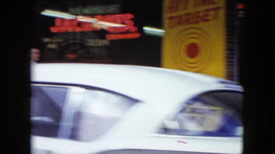 A white car moves across the foreground in a 1959 Nevada street scene with red neon and a yellow target-style sign behind it.