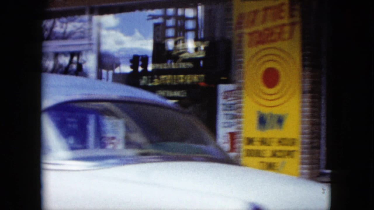 1959 Nevada street frame with a white classic car passing storefront windows and a yellow target-game poster on a brick building.