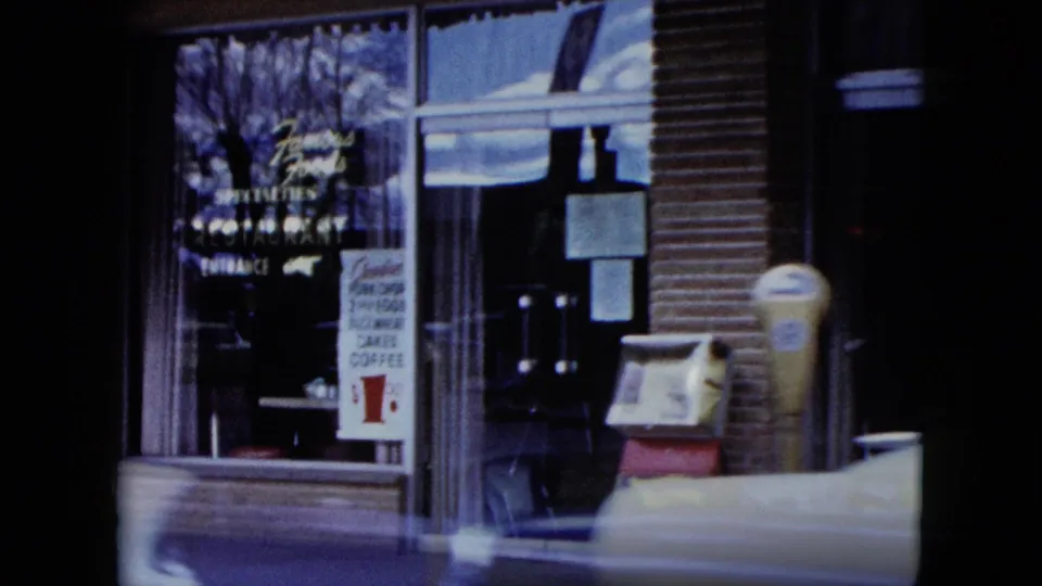 Nevada storefront view with window signs, a parking meter, and a newspaper box beside the curb in grainy 8mm color.