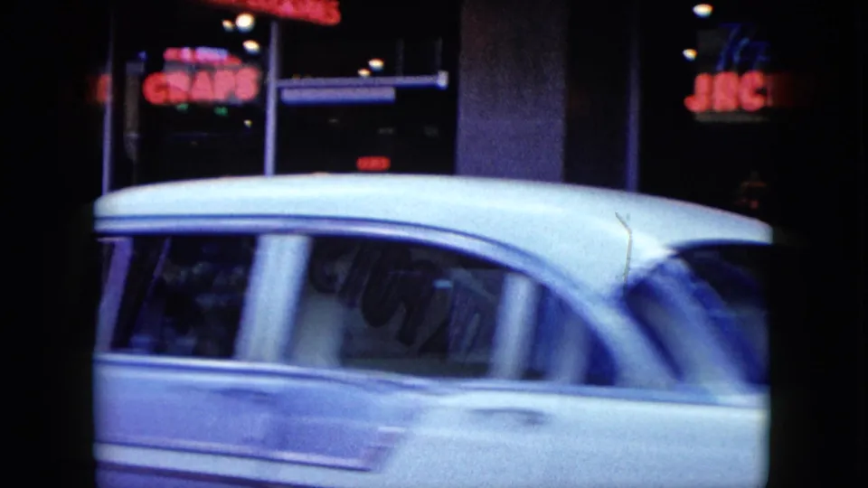 White mid-century sedan moving past red neon jackpot signage and dark storefront glass in 1959 Nevada street footage.