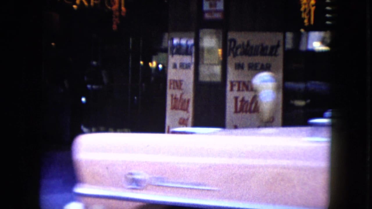 Rear of a pale classic car passing restaurant signs on a dark city street.