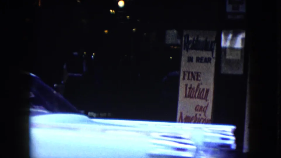 Night street view with a restaurant sign partly reading fine Italian and American food behind the bright hood of a passing car.