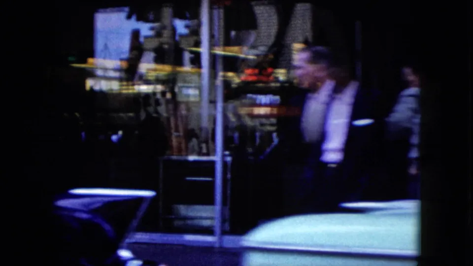 Two visible pedestrians stand or walk near a lit storefront window while classic cars pass during a 1959 Nevada night drive.
