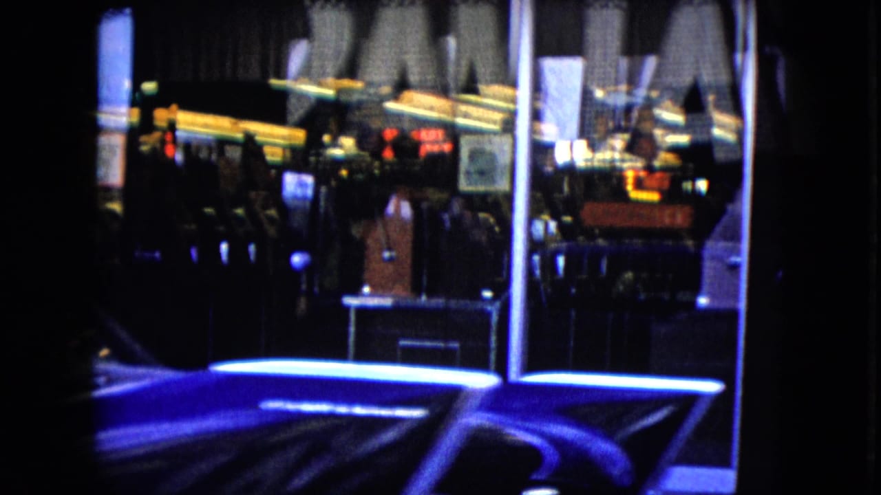 1959 Nevada home-movie frame showing a nighttime downtown storefront with reflected neon, dark sidewalk figures, and a shiny blue car moving across the foreground.