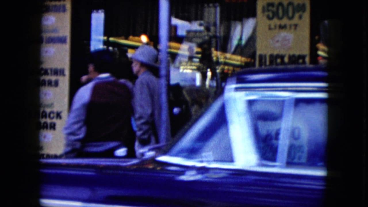 Two men on a sidewalk near casino signage as a blue car passes across the lower frame.