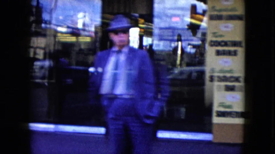 1959 Nevada home-movie frame showing a suited man in a brimmed hat walking past a neon-lit storefront with gaming or lounge signage and street reflections.