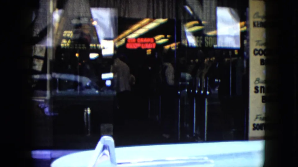 1959 Nevada nighttime street frame with neon cocktail lounge signage, dark glass doors, a pedestrian silhouette, and a pale car hood crossing the foreground.