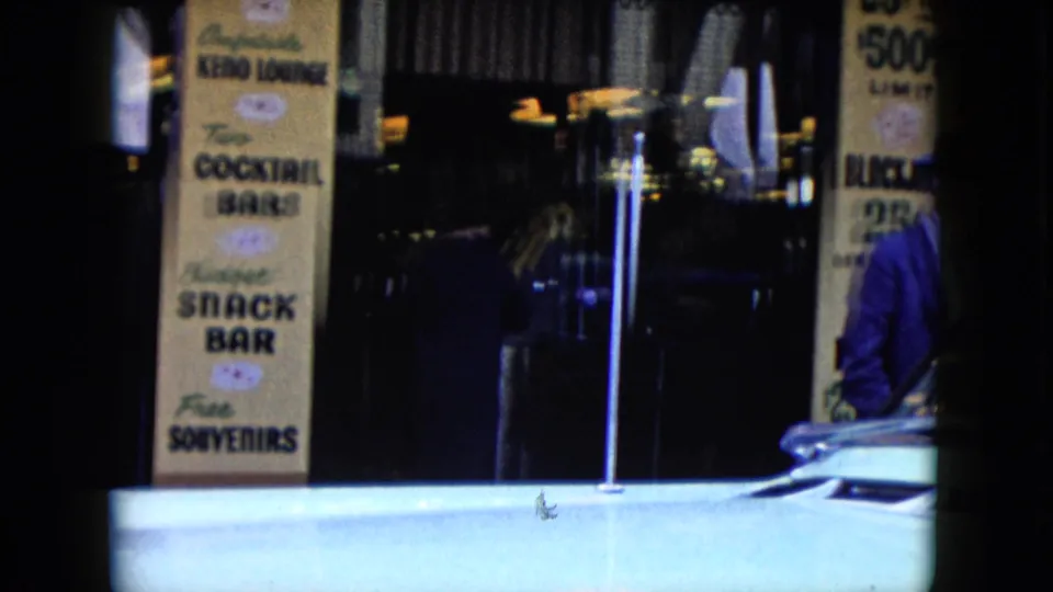 1959 Nevada storefront detail showing Keno lounge, cocktail bar, snack bar, and souvenir text with a light-colored classic car crossing the lower frame.