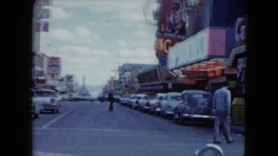 Wide Nevada street view from supplied 1959 metadata with rows of parked cars, pedestrians, storefront signage, and a distant tower-like structure.