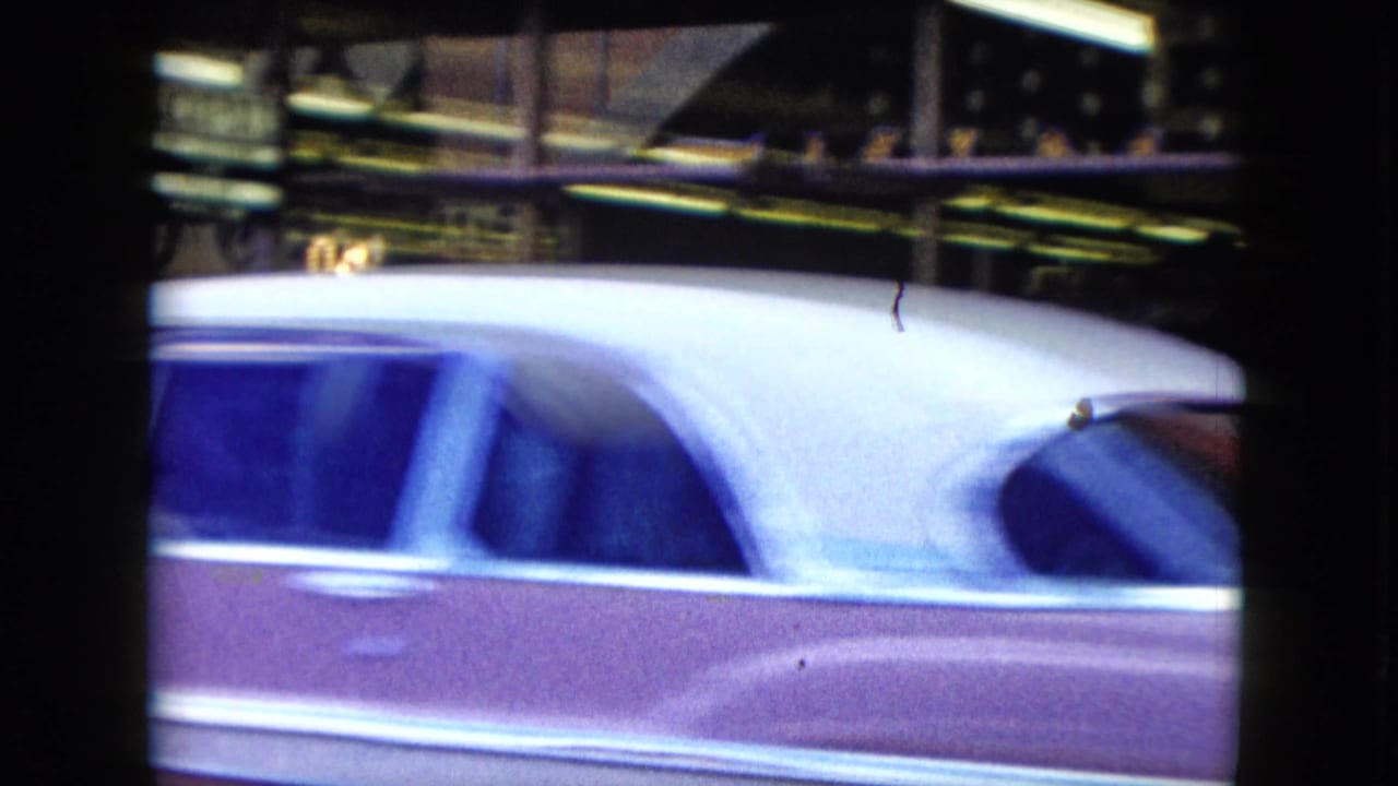 A mid-century car with a taxi-style roof sign passes across the frame, partially obscuring the storefronts behind it.