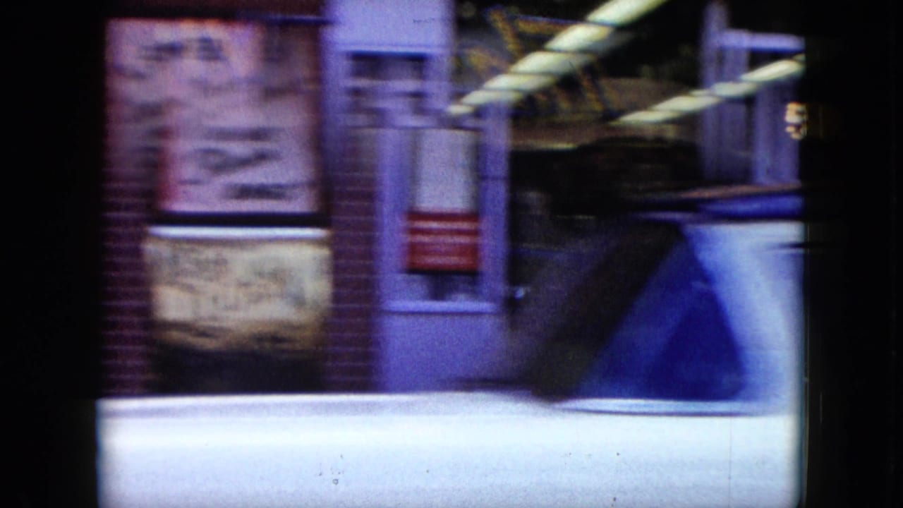 A white sedan passes in front of storefronts and signs.