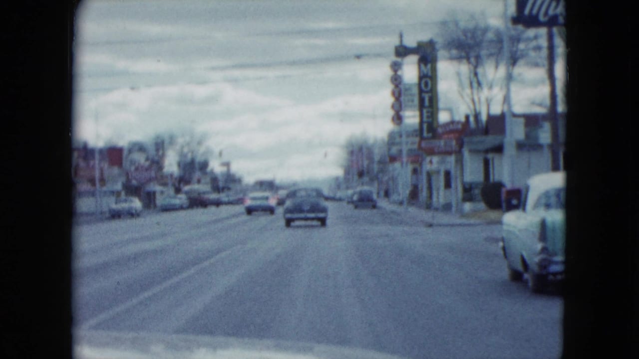1959 Nevada home-movie frame looking down a road past period cars, a vertical motel sign, low businesses, and bare trees.