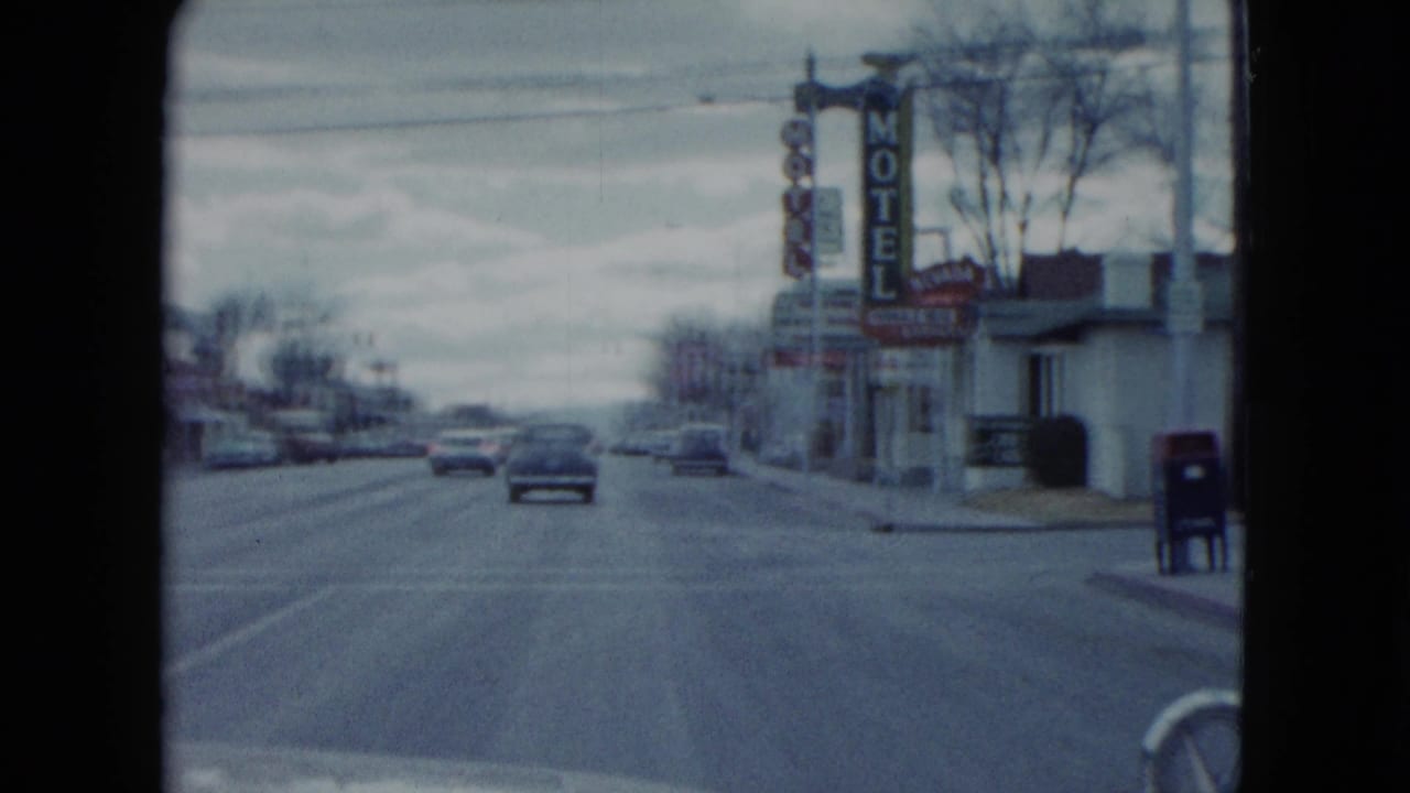 Roadway seen from a moving car with traffic ahead, motel signs, storefronts, and a mailbox or bin at right.