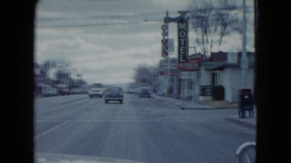 1959 Nevada street view from a moving vehicle, with cars ahead, motel signage, roadside buildings, and a dark curbside box.