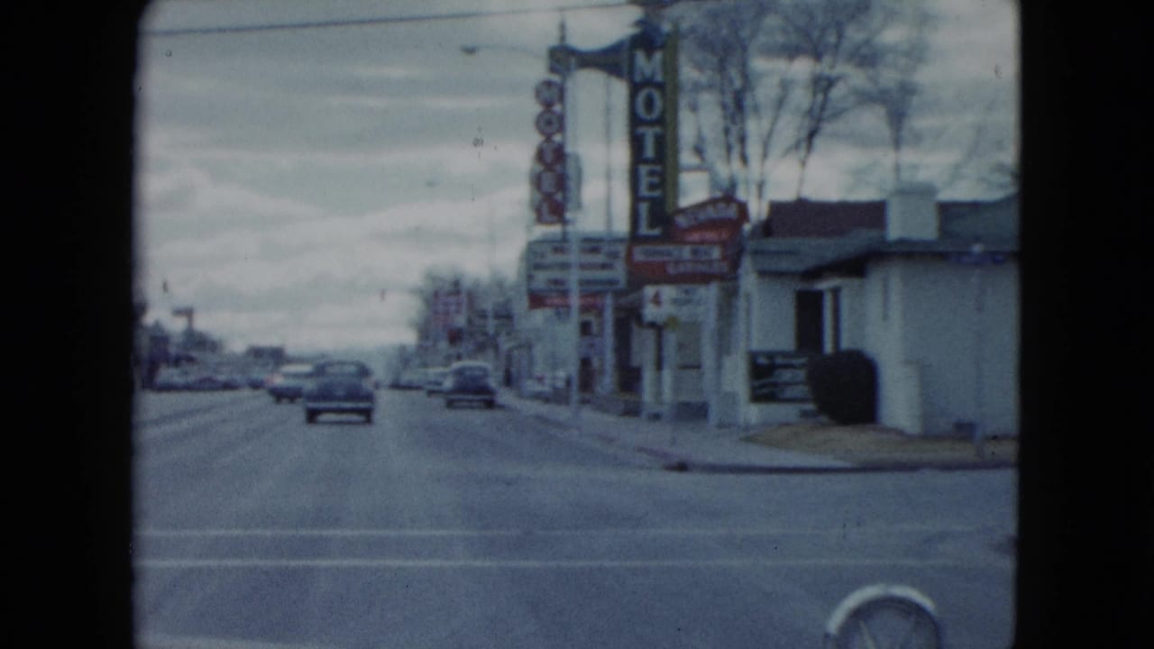 1959 Nevada roadside frame showing a prominent vertical motel sign, cars on the road, low commercial buildings, and sidewalks.